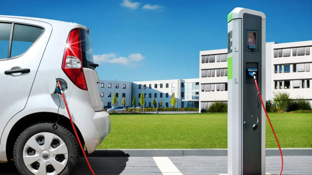 Auto bianca in carica presso una colonnina, pronta per la ricarica per auto elettriche. Edificio moderno sullo sfondo.