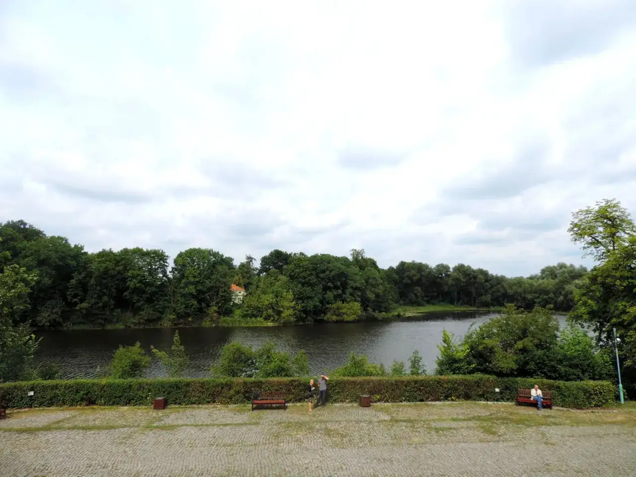 Spokojne jezioro Rokola otoczone bujną zielenią drzew. Na pierwszym planie brukowany plac z ławkami, gdzie odpoczywają ludzie.
