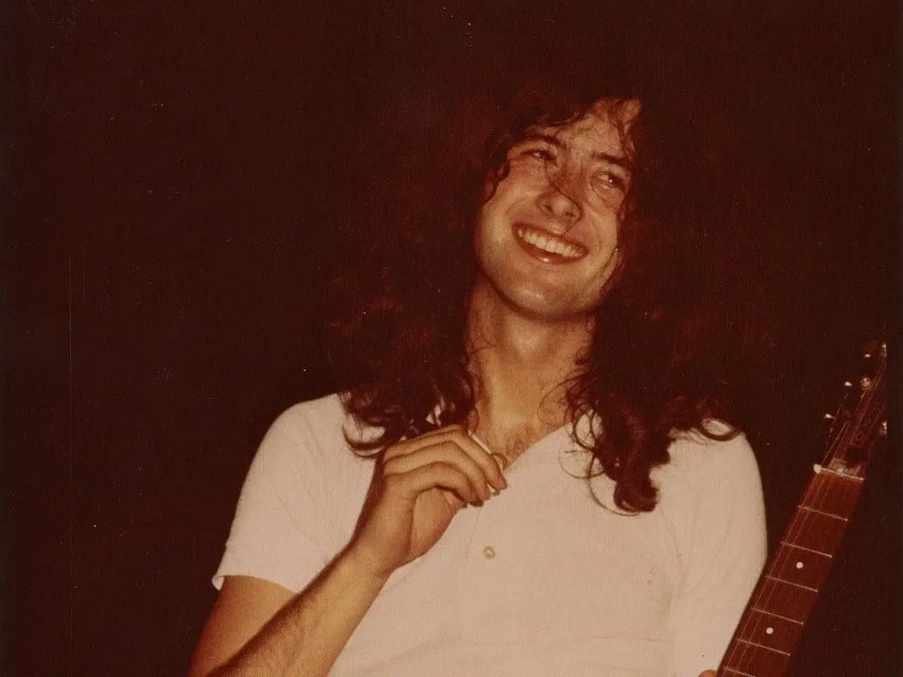 Uśmiechnięty Jimmy Page, gitarzysta zespołu Led Zeppelin, z burzą włosów i w białej koszulce, gotowy do gry.