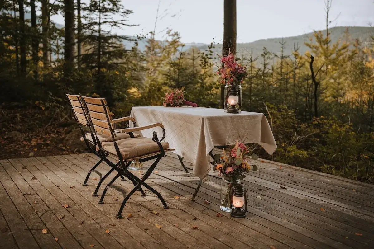 Zwei Stühle und ein Tisch mit Blumen und Laternen auf einer Holzterrasse im Wald. Ein romantischer Ort, um heimlich heiraten zu wollen.