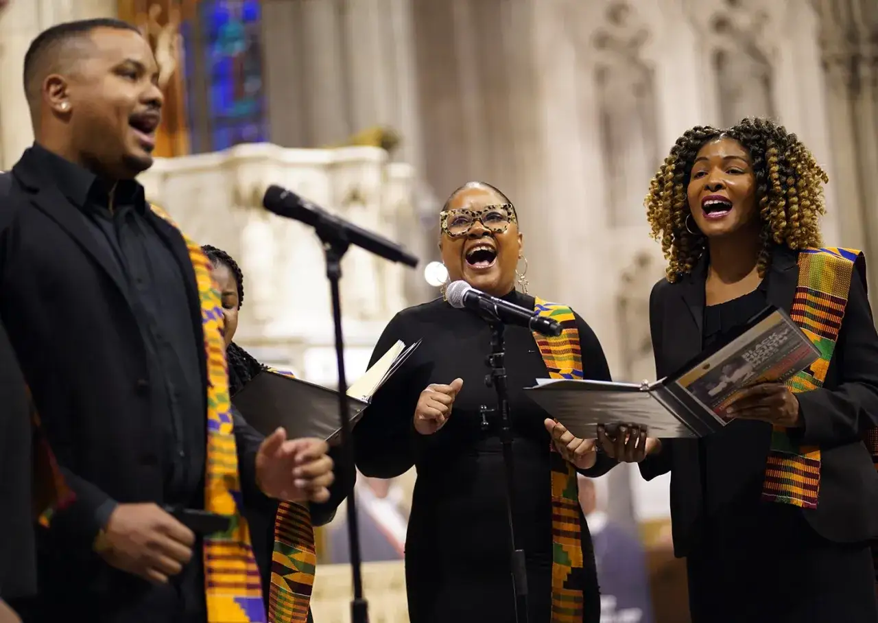 A choir sings with passion, their voices rising in powerful chant ideas. They wear vibrant Kente cloth sashes over black attire, holding sheet music.