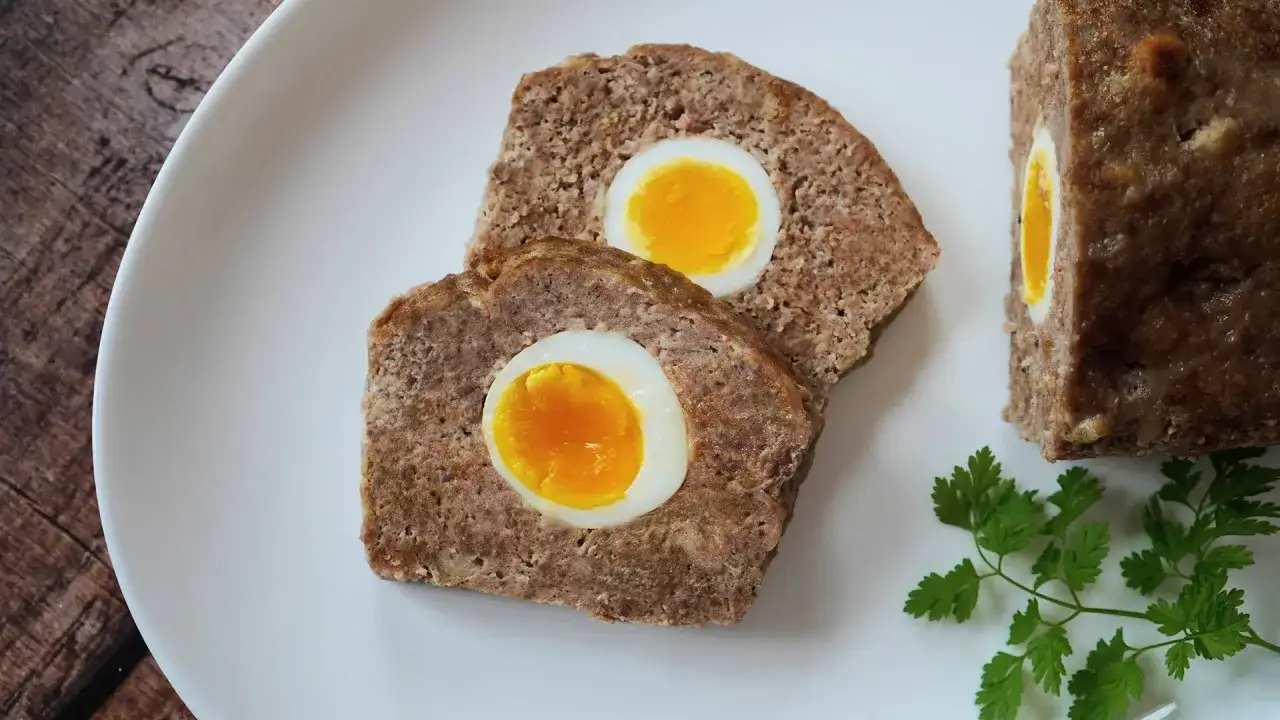Zwei Scheiben falscher Hase mit gekochtem Ei im Inneren, daneben ein Stück und Petersilie.