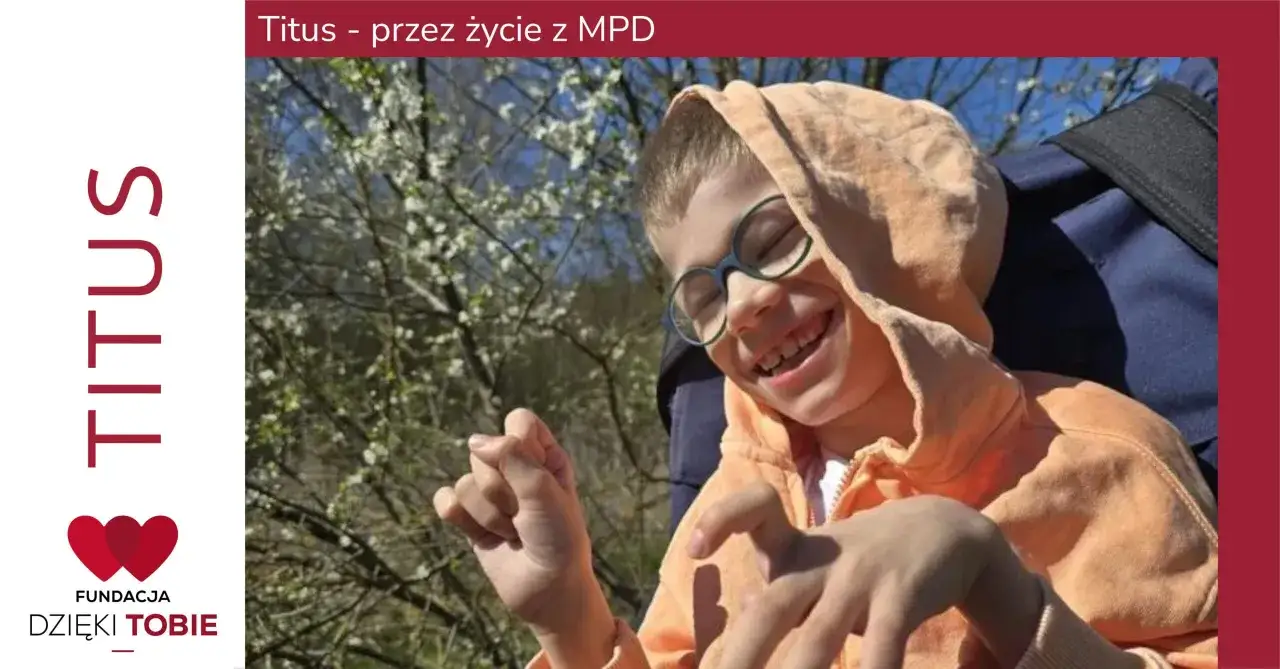 Uśmiechnięty chłopiec w pomarańczowej bluzie z kapturem, w okularach, siedzi na wózku. Jego dłonie są lekko zgięte, co może być związane z tetraplegią.