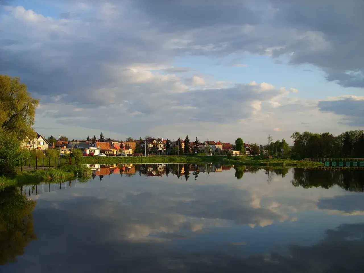Spokojne jezioro odbija zabudowania i chmury. Kazimierza Wielka atrakcje to m.in. malownicze widoki nad wodą.