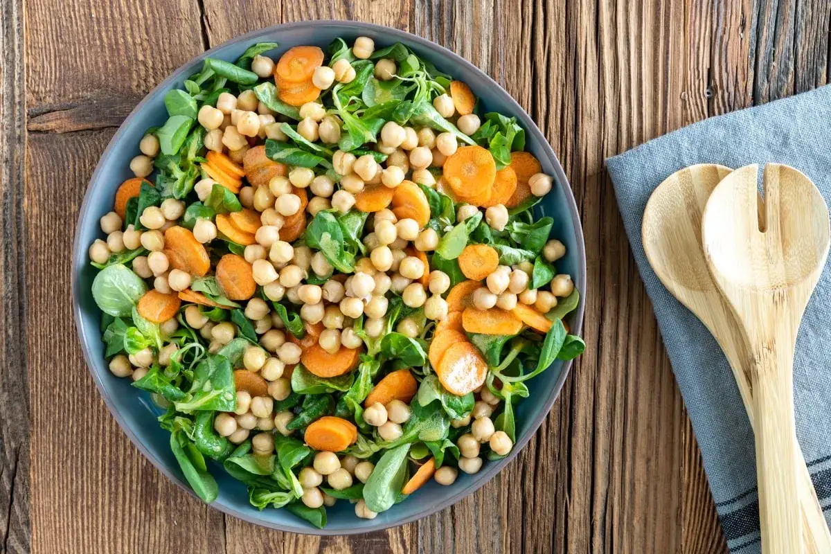 Un'insalata di ceci fresca e colorata, con carote a rondelle e verdure a foglia verde, servita in un piatto blu su un tavolo di legno.