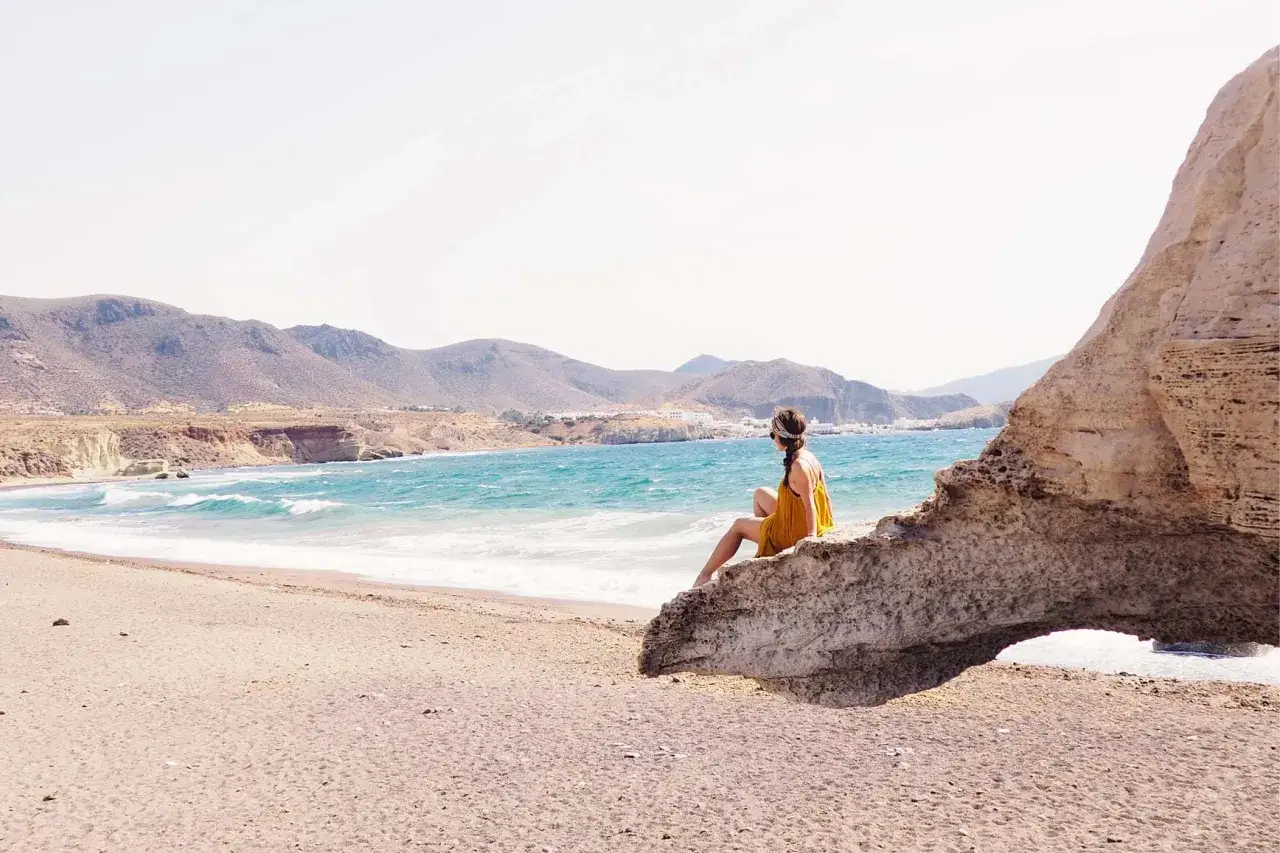 Mujer sentada en roca, contemplando las **playas de Cabo de Gata** y el mar turquesa. Paisaje árido y montañoso al fondo.