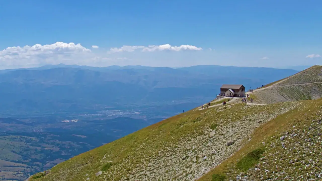 Il **Rifugio Duca degli Abruzzi** domina un paesaggio montano mozzafiato, con un villaggio in lontananza sotto un cielo azzurro.