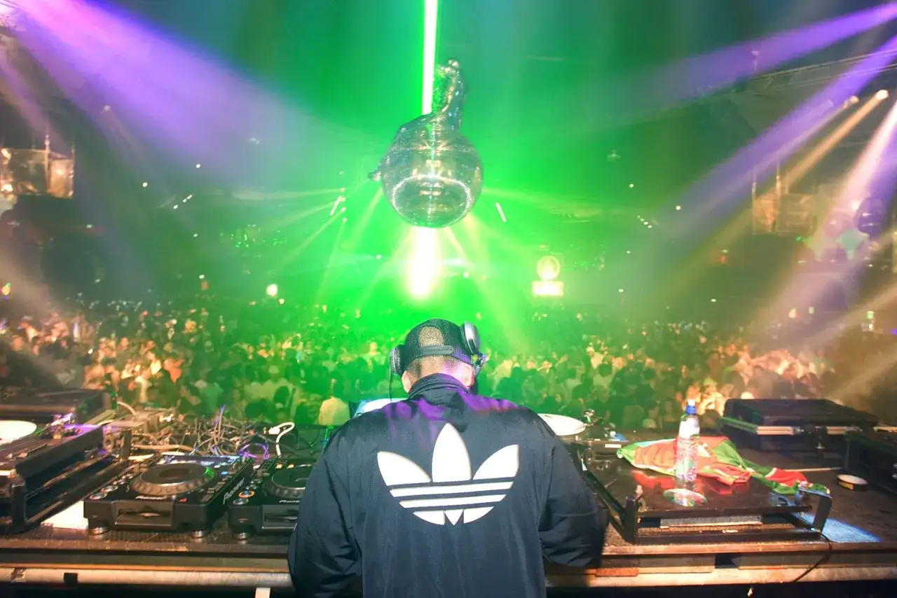 DJ w czapce i słuchawkach gra na konsoli, tłum szaleje w rytm techno przy zielonych i fioletowych światłach.