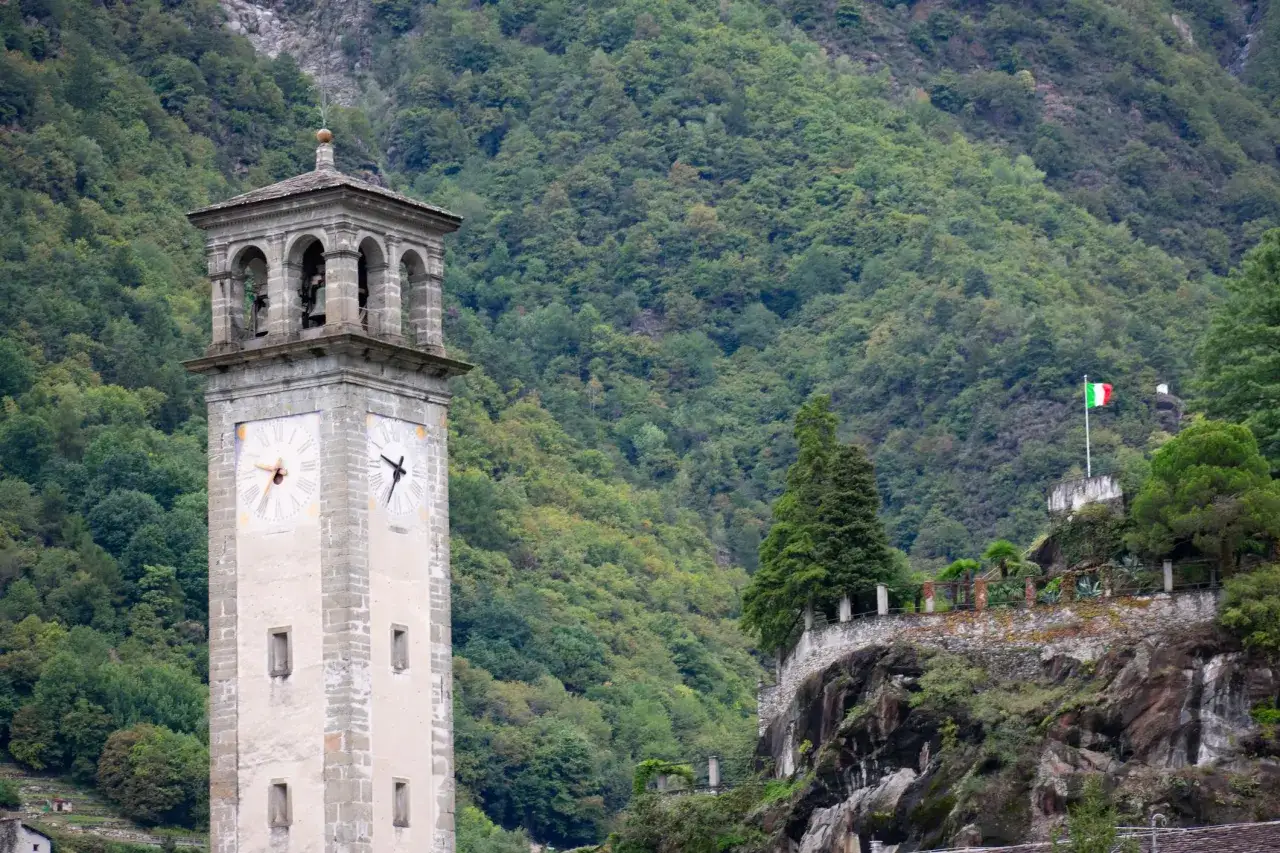 Torre campanaria con orologio a Chiavenna, cosa vedere: un paesaggio montano lussureggiante e la bandiera italiana sventolano su una roccia.