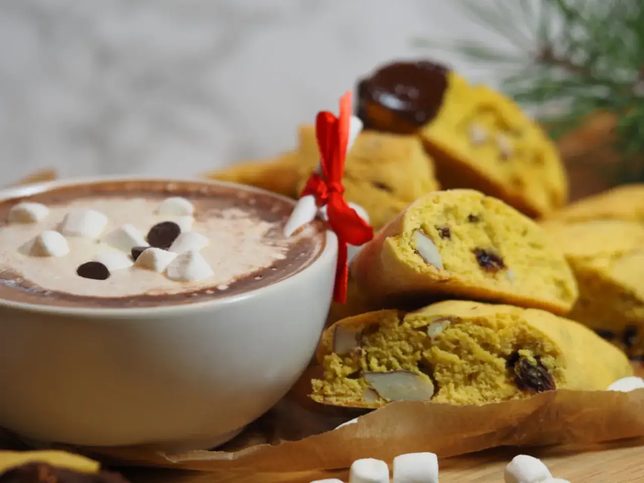 En kopp varm choklad med marshmallows och chokladbitar bredvid en h&ouml;g med saffransbiscotti med mandel, bundna med ett r&ouml;tt band.