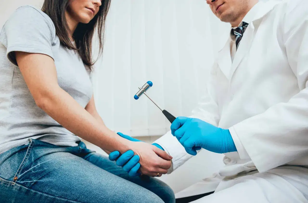 Lekarz w niebieskich rękawiczkach wykonuje próbę Romberga u pacjentki, używając młoteczka neurologicznego.