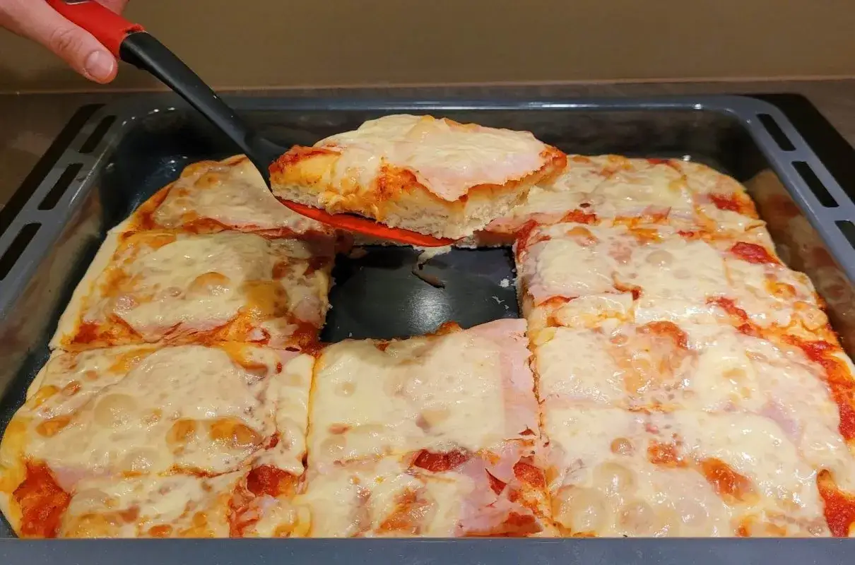 Kawałek pysznej, domowej pizzy na grubym cieście, z ciągnącym się serem i szynką, właśnie jest wyjmowany z blachy.