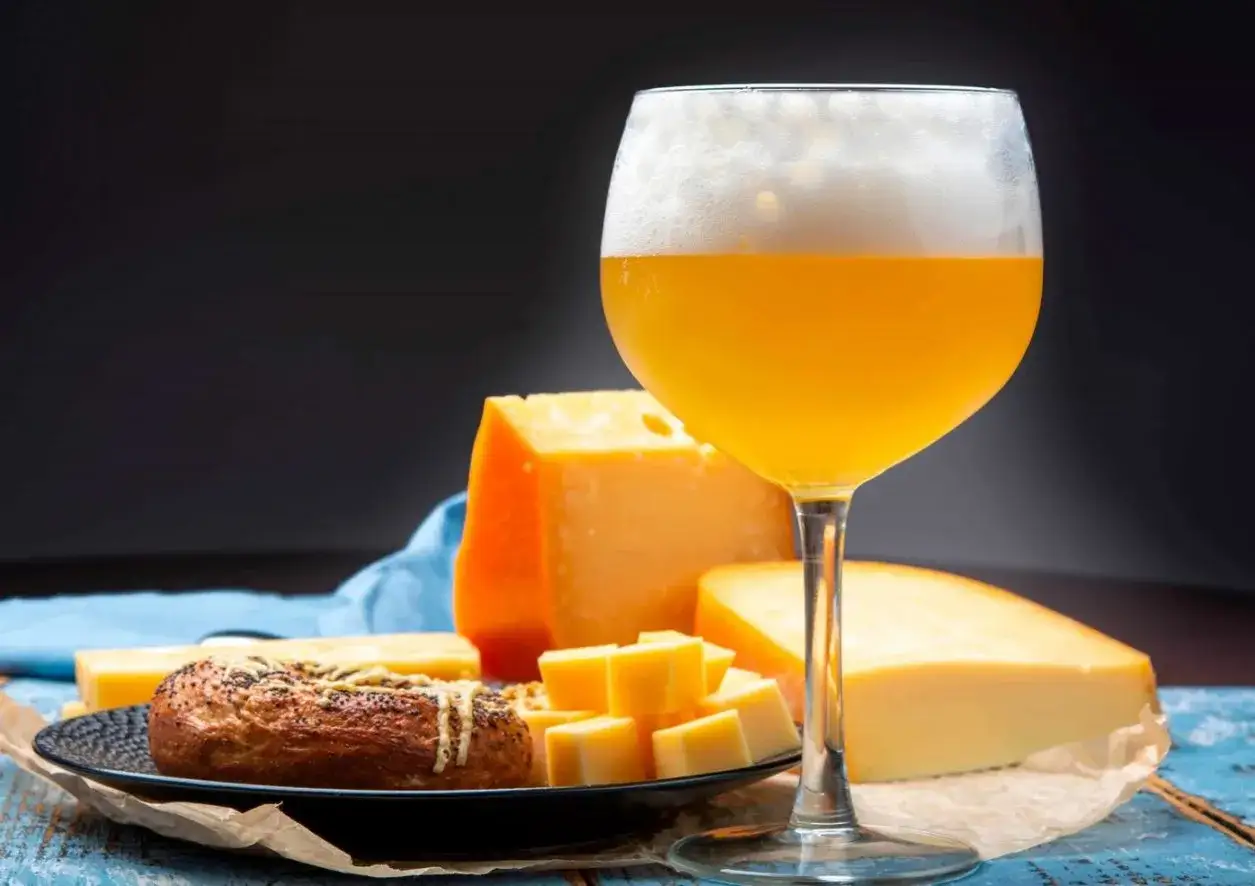 Un bicchiere di birra dorata con schiuma abbondante, accanto a vari formaggi stagionati e un panino con semi.