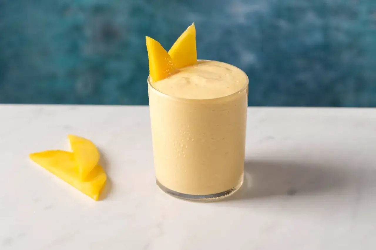 En kr&auml;mig mango lassi i ett glas, dekorerad med mangoslantar. Bredvid ligger ytterligare tv&aring; mangoslantar p&aring; en marmoryta.