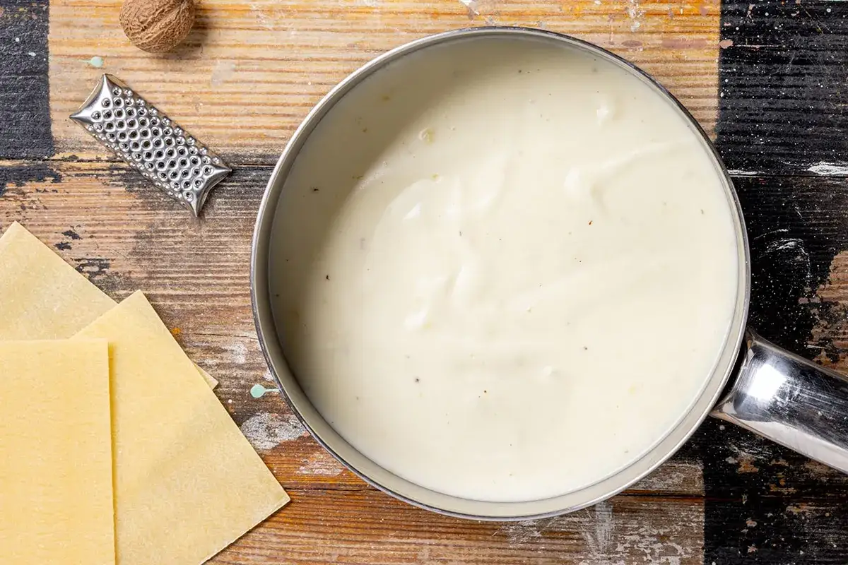 Besciamella senza burro cremosa in pentola, con sfoglie di pasta per lasagne, noce moscata e grattugia.