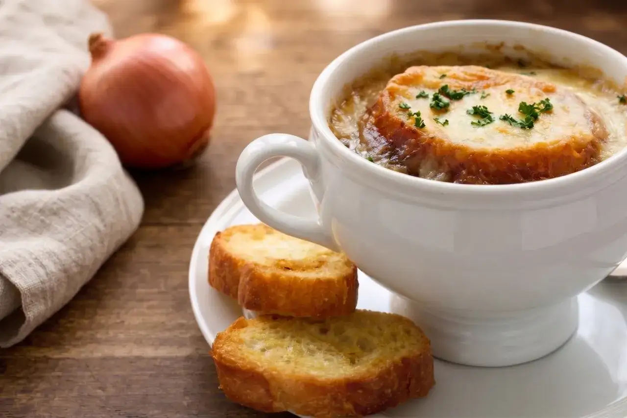 Una tazza di zuppa di cipolle fumante, con crostini dorati e formaggio fuso, accanto a una cipolla cruda e un tovagliolo di lino.