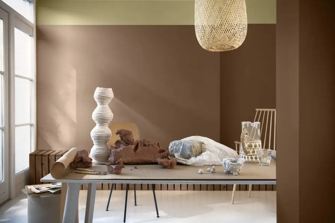 Un salon aux murs beige et marron chocolat, avec une table de potier recouverte d'argile et d'outils.
