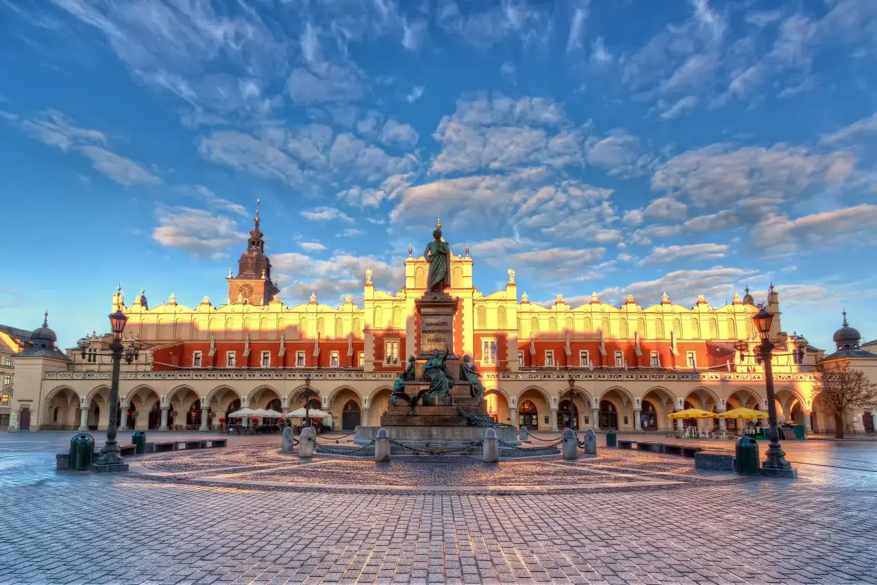 Rynek Główny Kraków o wschodzie słońca. Monumentalny Sukiennice i pomnik Mickiewicza na tle błękitnego nieba z chmurami.