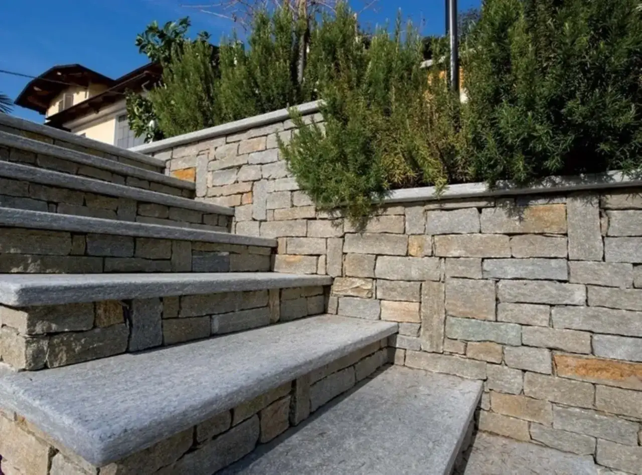 Scale esterne in muratura con gradini in pietra grigia, circondate da un muro di pietra grezza e piante verdi rigogliose.