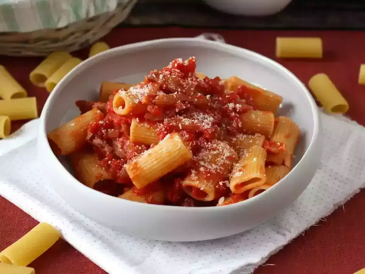 Pyszne rigatoni amatriciana w białej misce, posypane parmezanem. Obok leżą surowe rurki makaronu.