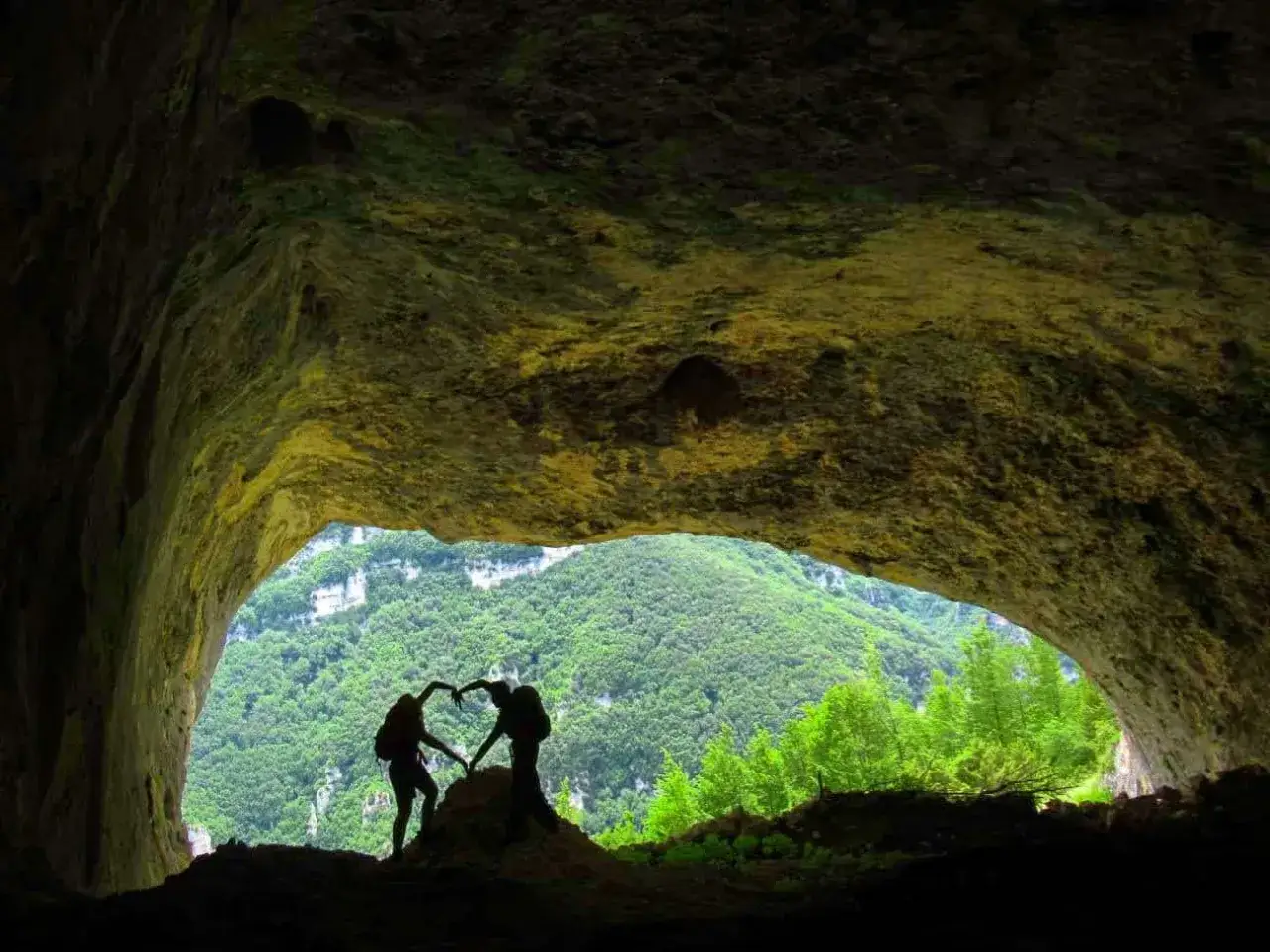 Coppia abbracciata a forma di cuore all'ingresso di una grotta nera, con vista su un paesaggio verdeggiante.
