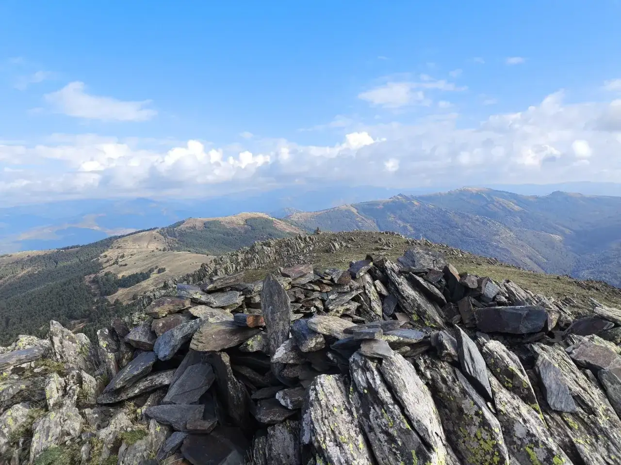 C&uacute;spide rocosa con un cairn, como si fueran las paredes de Buitrago, bajo un cielo azul con nubes. Paisaje monta&ntilde;oso al fondo.