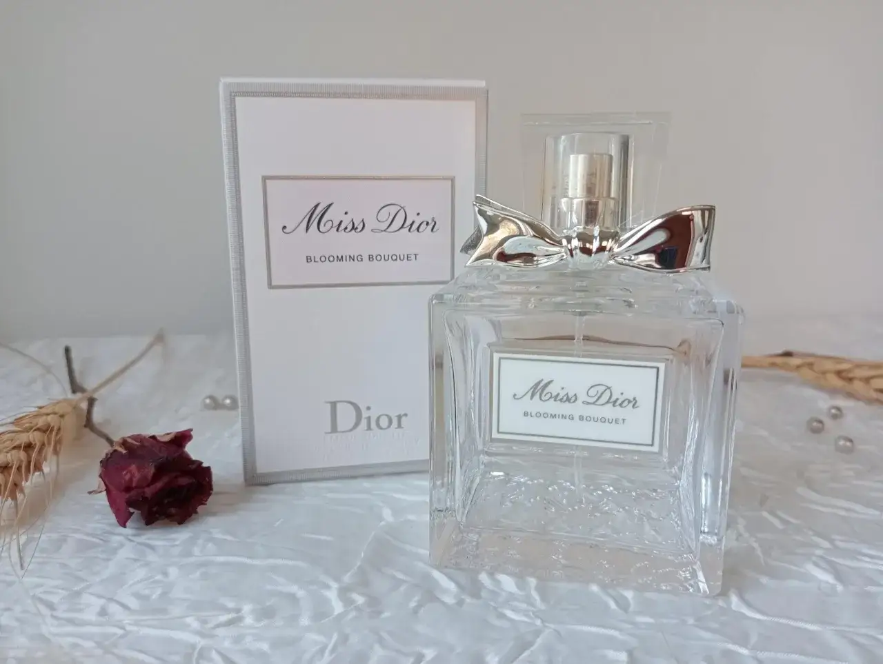 Elegancka butelka perfum Miss Dior Blooming Bouquet z ozdobną kokardką, obok pudełka i zasuszonej róży.