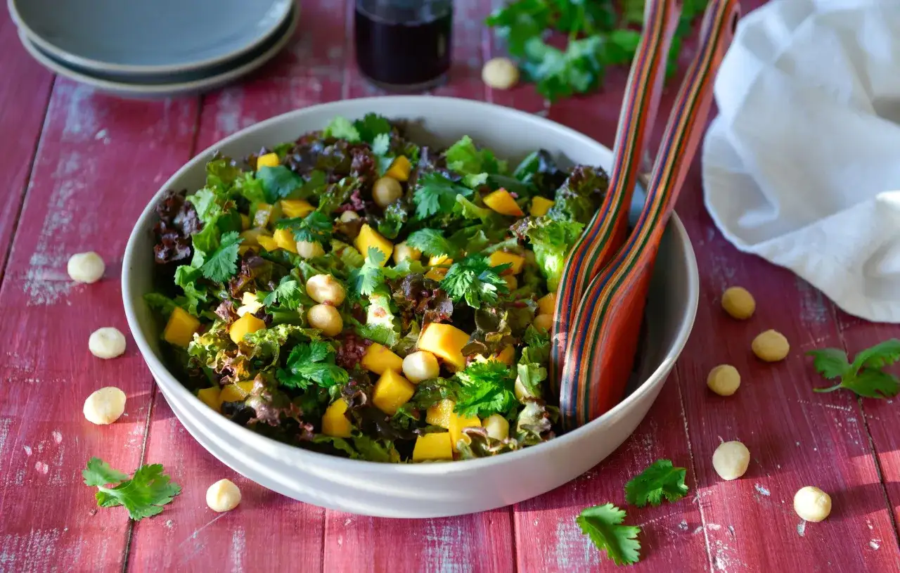 Bunter Mango Salat mit knackigen Macadamian&uuml;ssen und frischem Koriander, serviert in einer Sch&uuml;ssel auf einem roten Holztisch.