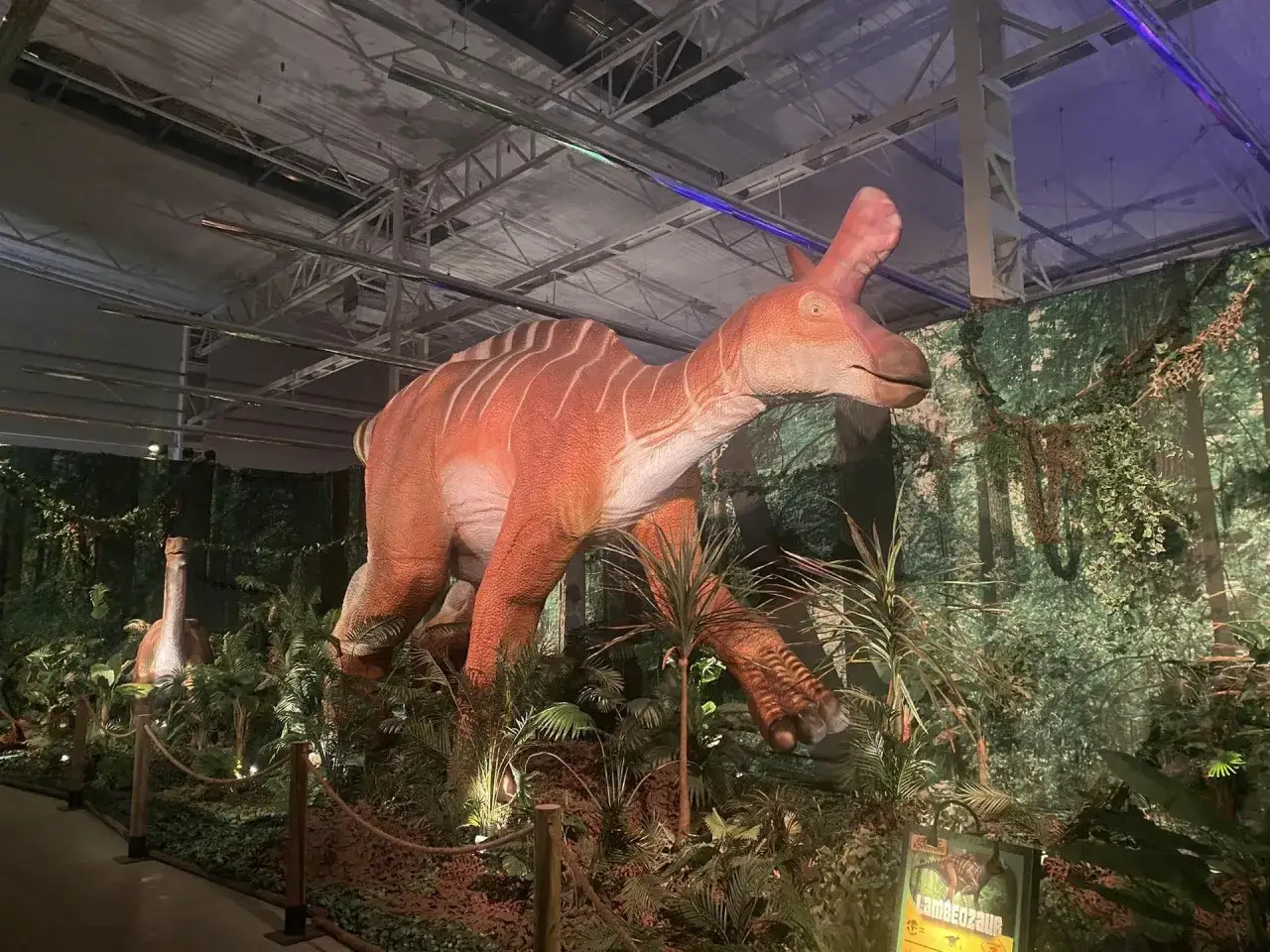 Wielki, pomarańczowy dinozaur z białymi pasami w otoczeniu bujnej roślinności. To część wystawy w muzeum dinozaur&oacute;w w Warszawie.