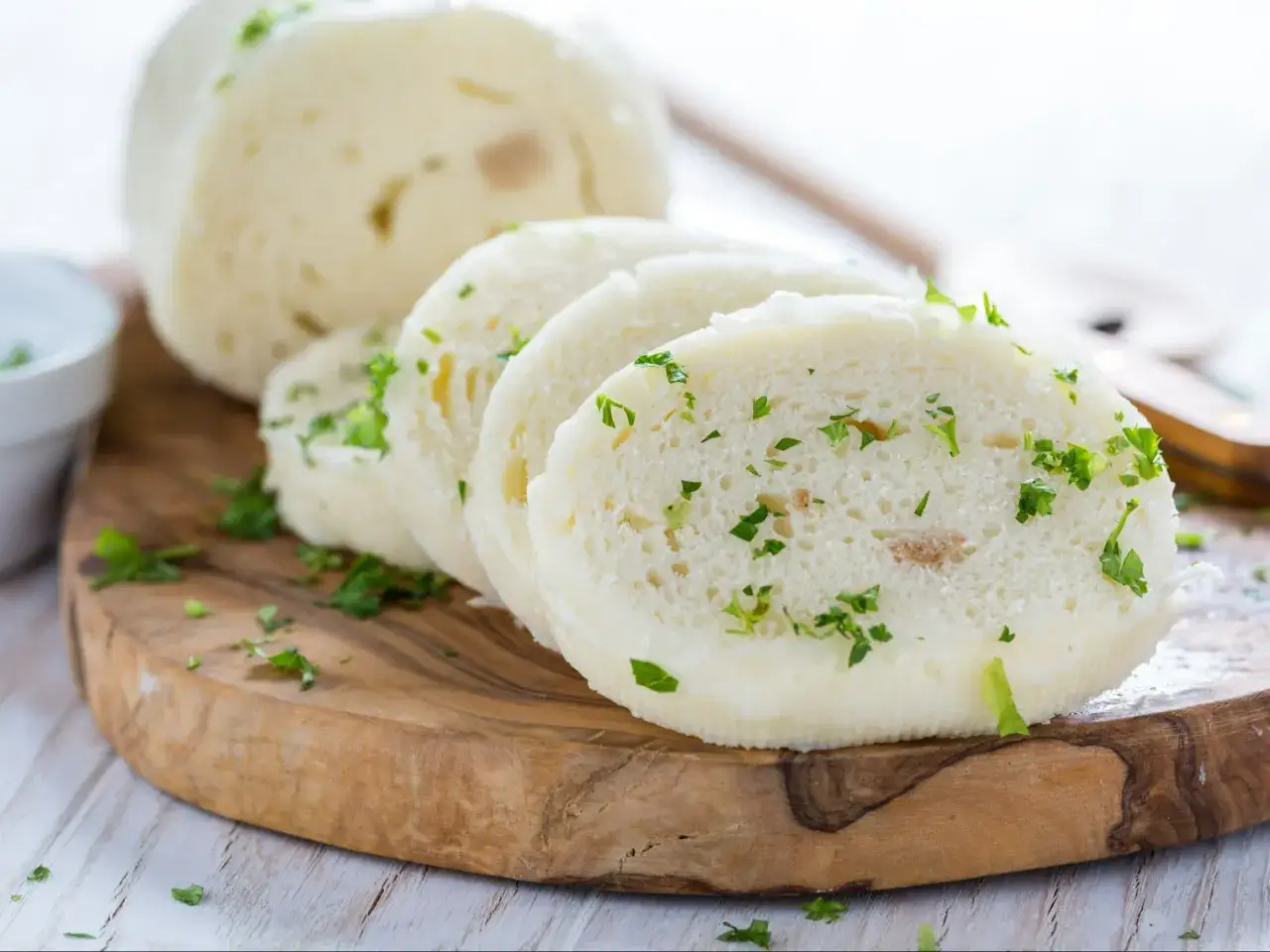 Knedle z chleba, pokrojone w plastry i posypane natką pietruszki, leżą na drewnianej desce.