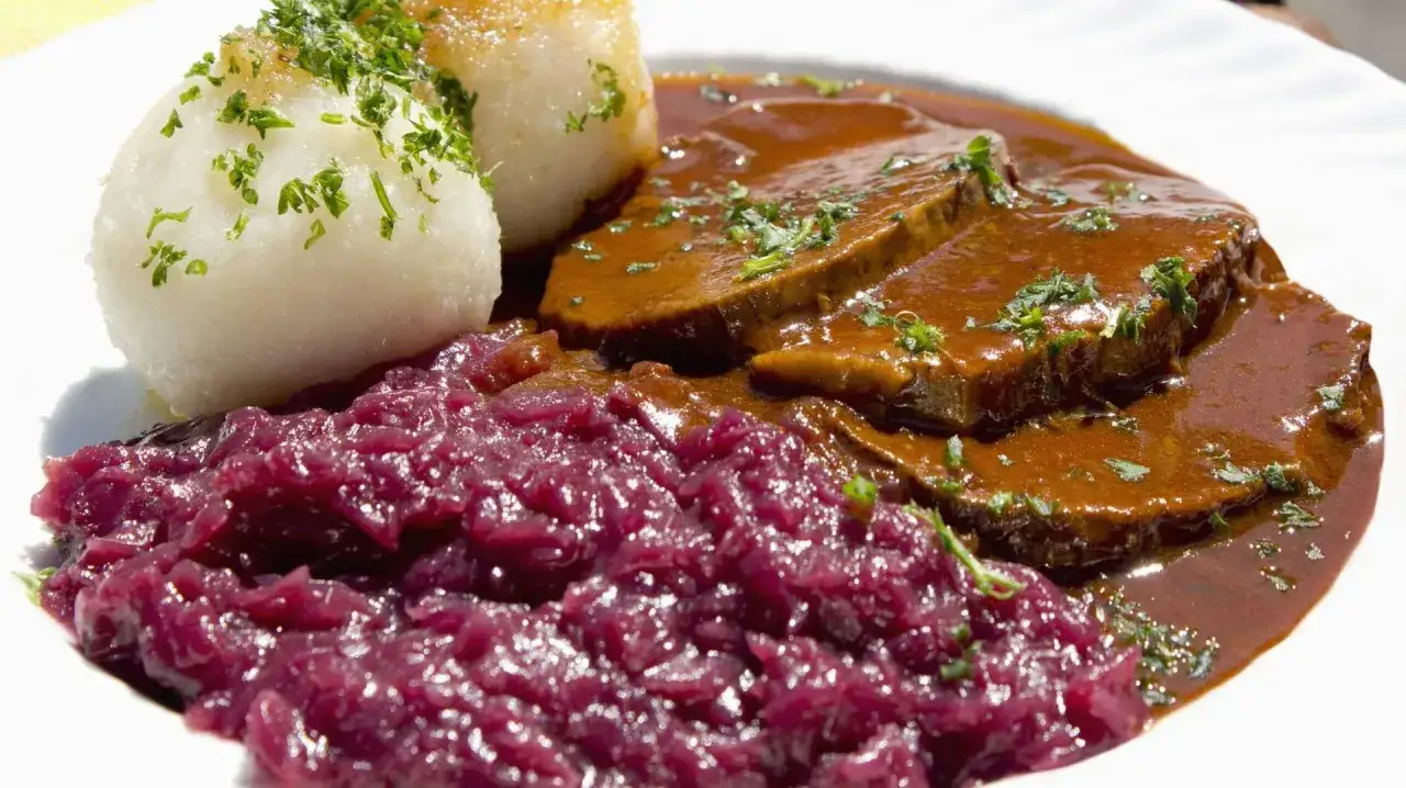 Ein Teller mit Scheiben von Sauerbraten in dunkler Soße, dazu Klöße und Rotkohl. Ein klassisches Sauerbraten Rezept.