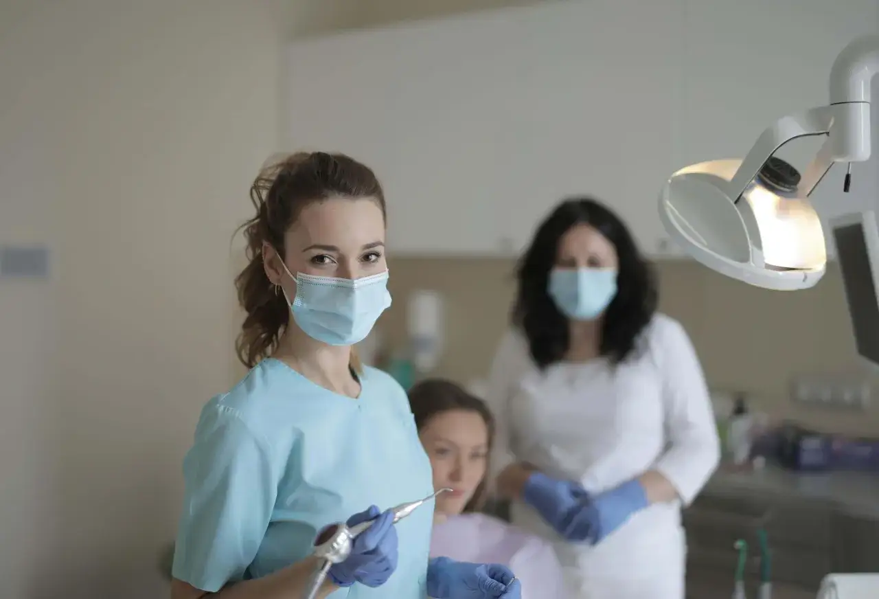 Dentystka w masce i rękawiczkach trzyma wiertło, gotowa do leczenia pacjenta. W tle druga lekarka i pacjentka, być może zmagająca się z abrazją zębów.