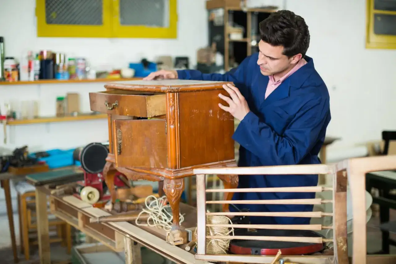 Un artisan examine un meuble en bois, prêt à le repeindre. Des outils et des matériaux sont visibles dans l'atelier.