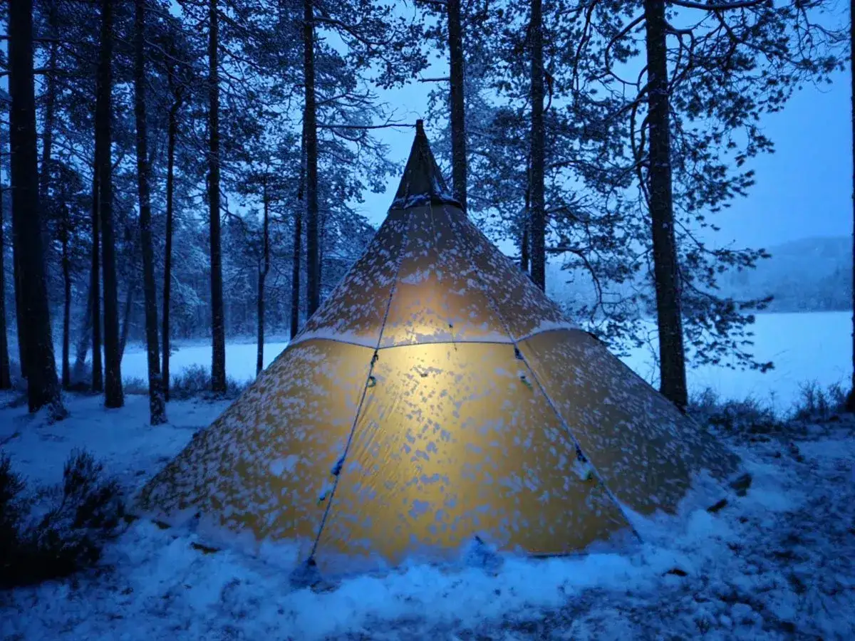 Varmt ljus lyser från ett snötäckt tält i en vinterlig skog. En mysig plats att sova ute på vintern.