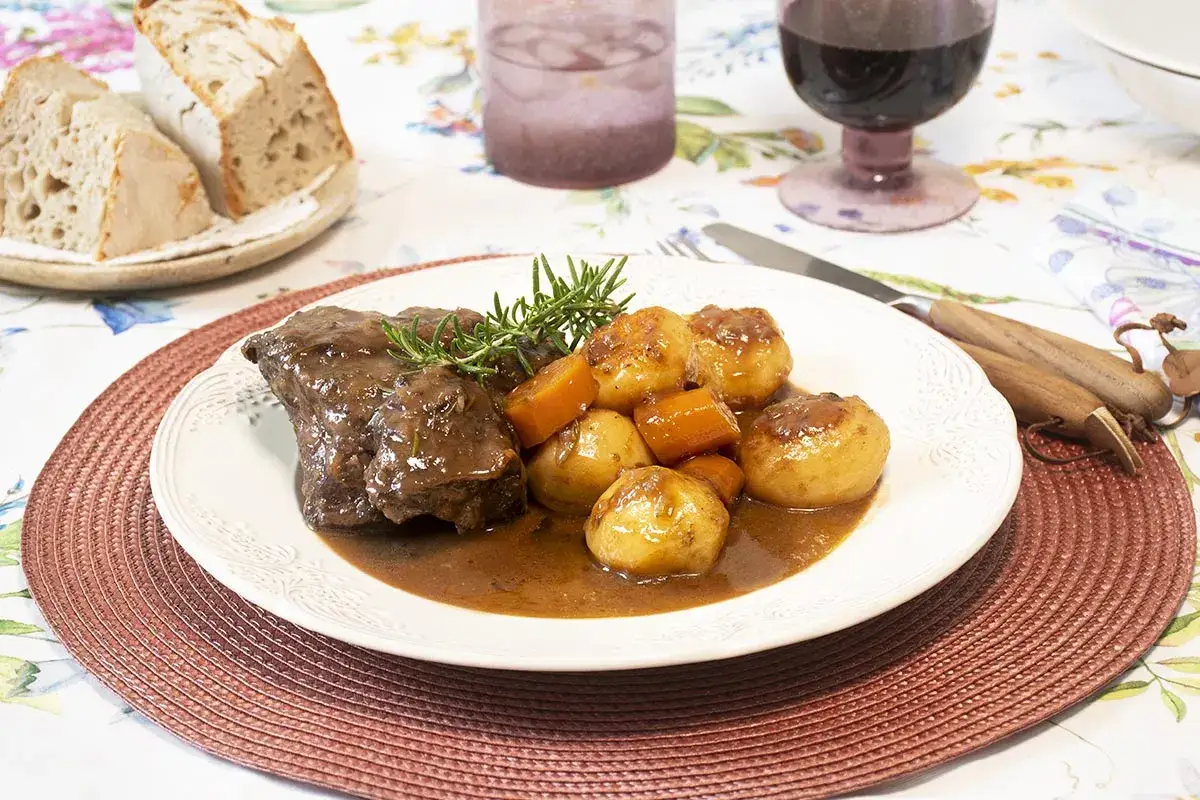 Carrilleras de ternera estofadas con patatas y zanahorias, servidas con pan r&uacute;stico y vino tinto.