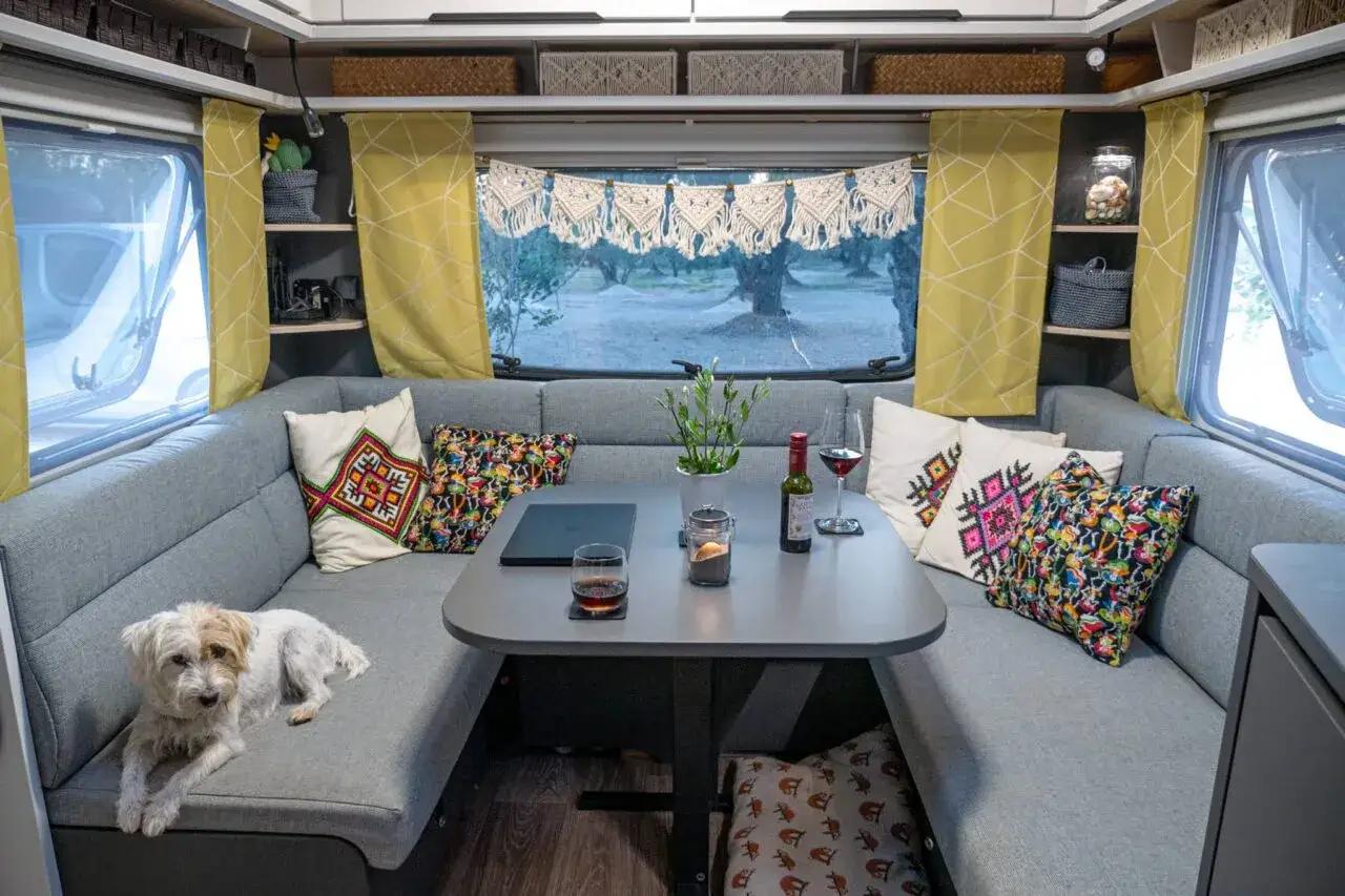 Gem&uuml;tliche Wohnwagen-Inneneinrichtung mit U-f&ouml;rmiger Sitzbank, bunten Kissen, Tisch mit Wein und einem Hund. Tolle Ideen f&uuml;r Wohnwagen innen.