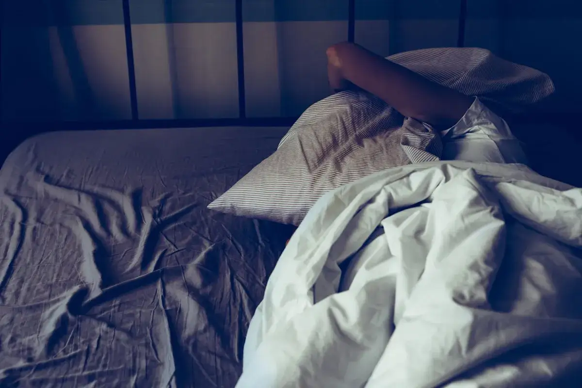 A person's arm is visible under rumpled sheets, suggesting insomnia before period. The bed is unmade, with a striped pillow.