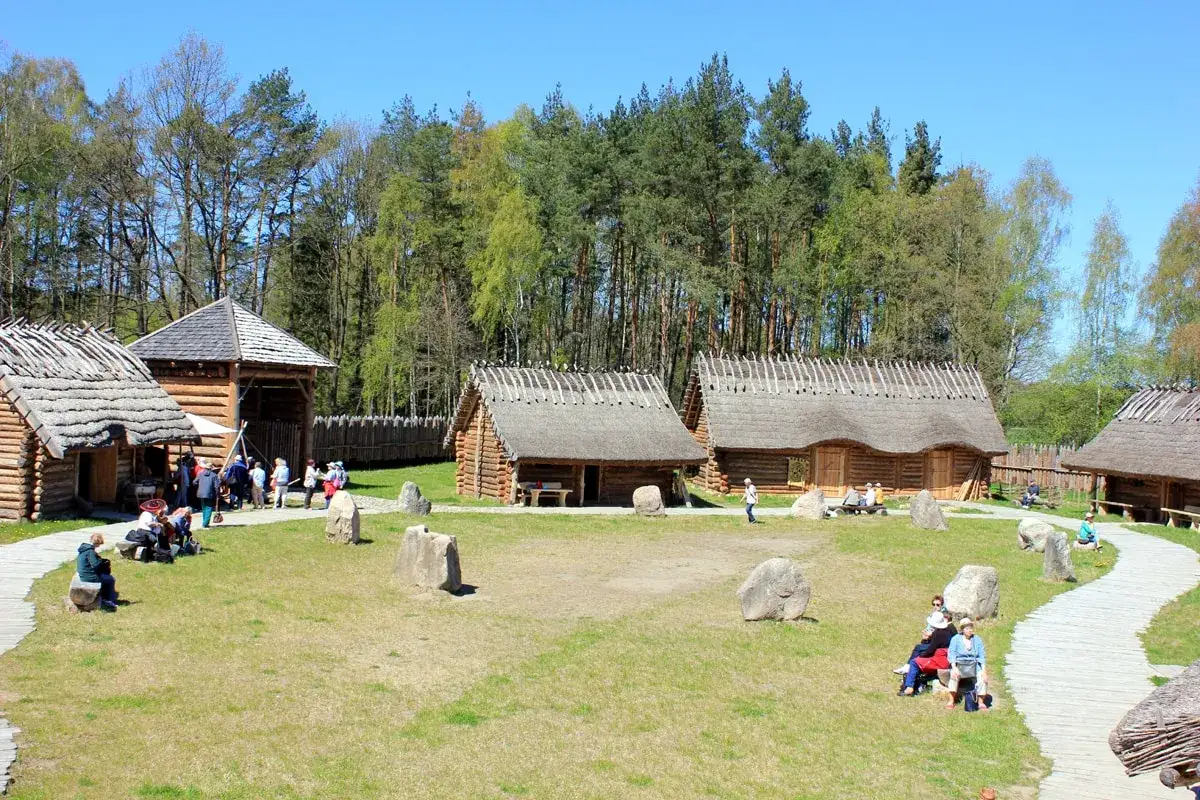 Odwiedzający zwiedzają osadę średniowieczną Sławutowo, podziwiając drewniane chaty kryte strzechą i bramę.