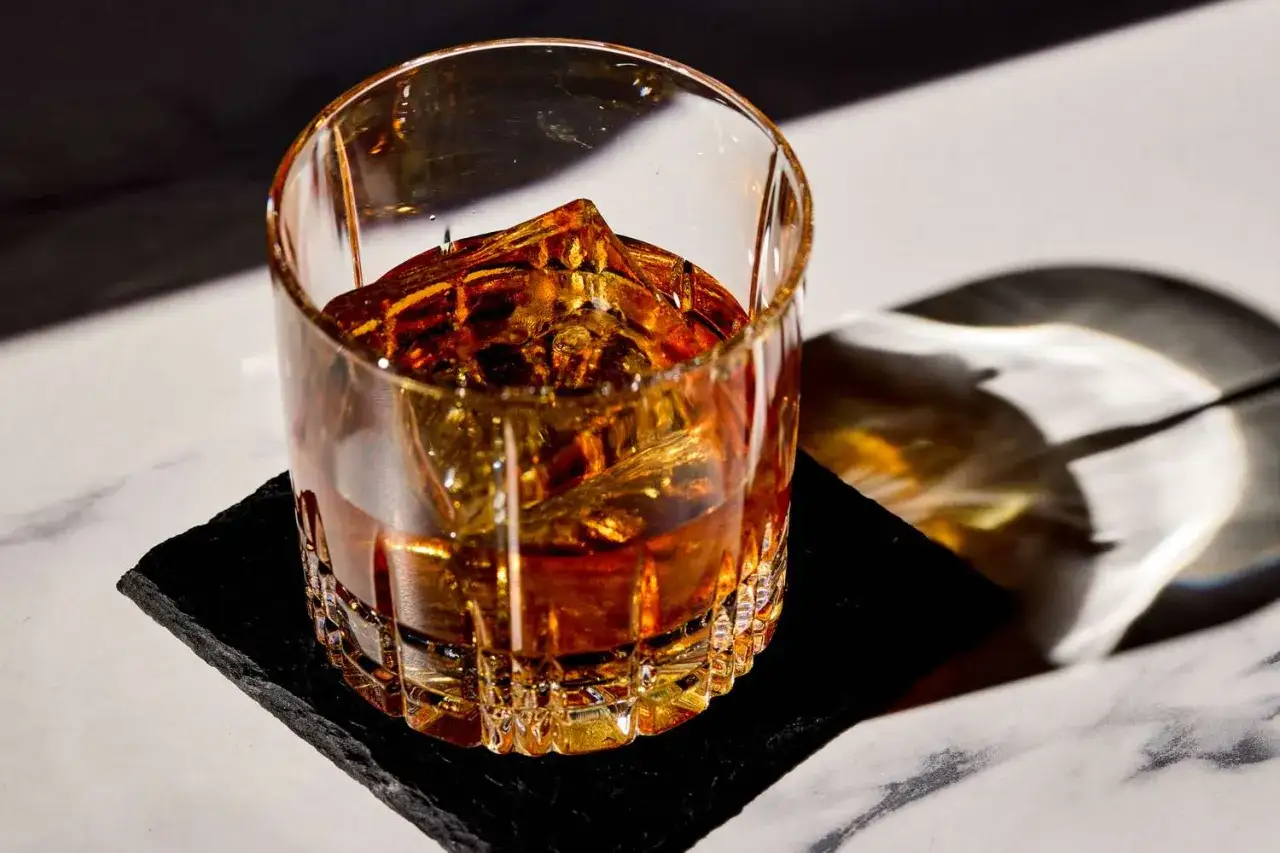 Ein bernsteinfarbener French Connection Cocktail mit Eisw&uuml;rfeln in einem geschliffenen Glas auf einem schwarzen Untersetzer.
