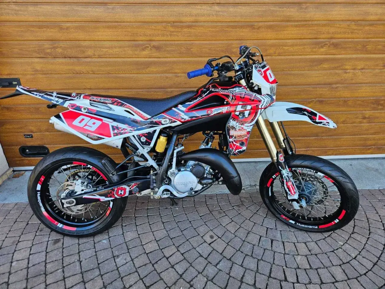 Husqvarna TE 125 z agresywnym oklejeniem w czerwono-białe wzory, gotowa do jazdy po torze.