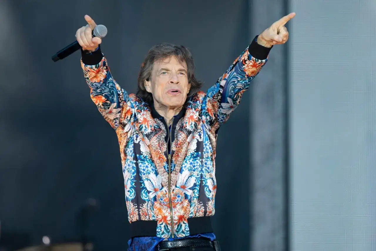 Mick Jagger z zespołu The Rolling Stones w kolorowej kurtce, z mikrofonem w dłoni, wskazuje palcem na publiczność.