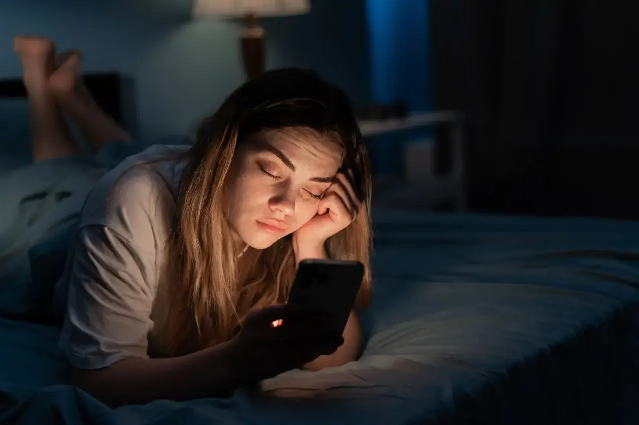 Zmęczona dziewczyna leży w ł&oacute;żku, przeglądając telefon. Czy to godziny szczytu na Tinderze, czy po prostu szuka czegoś do oglądania przed snem?