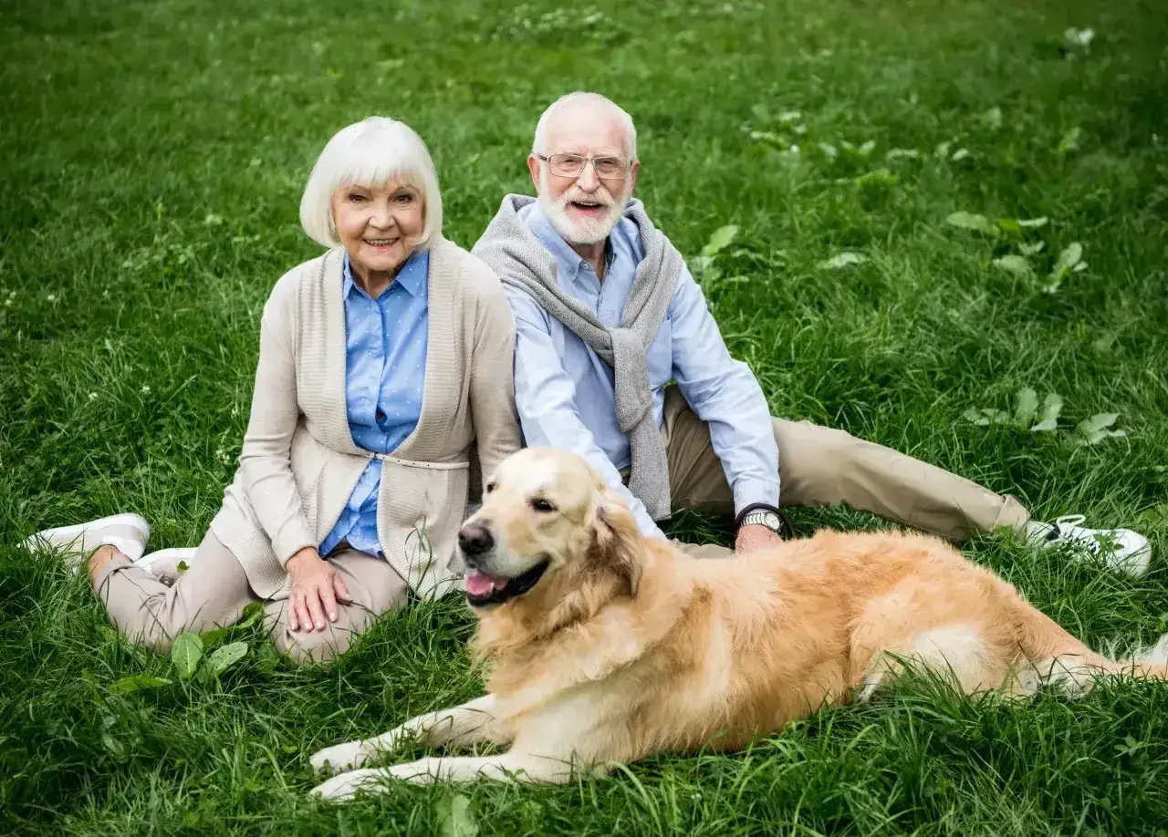 Uśmiechnięci seniorzy siedzą na trawie z psem. Złoty retriever leży obok, idealny pies dla seniora, który wnosi radość i towarzystwo.