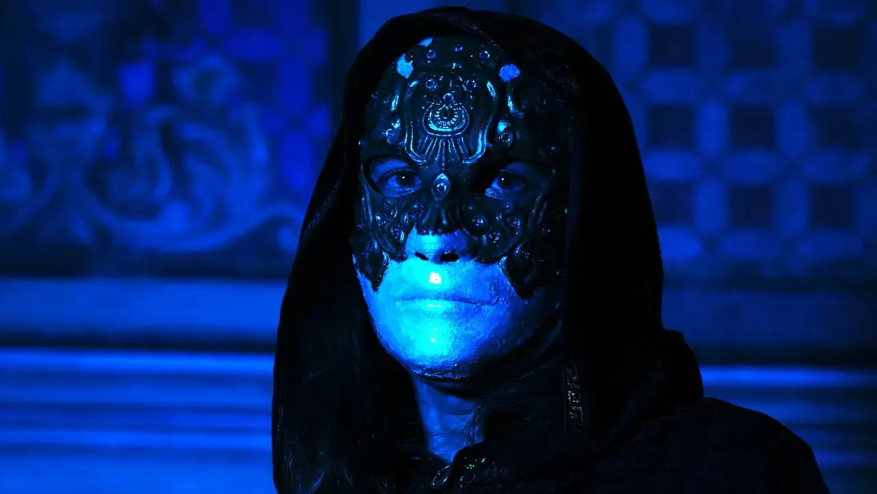 Volto coperto da maschera ornata, illuminato da luce blu. Un'espressione che evoca un profondo **horror psicologico**, tra mistero e inquietudine.