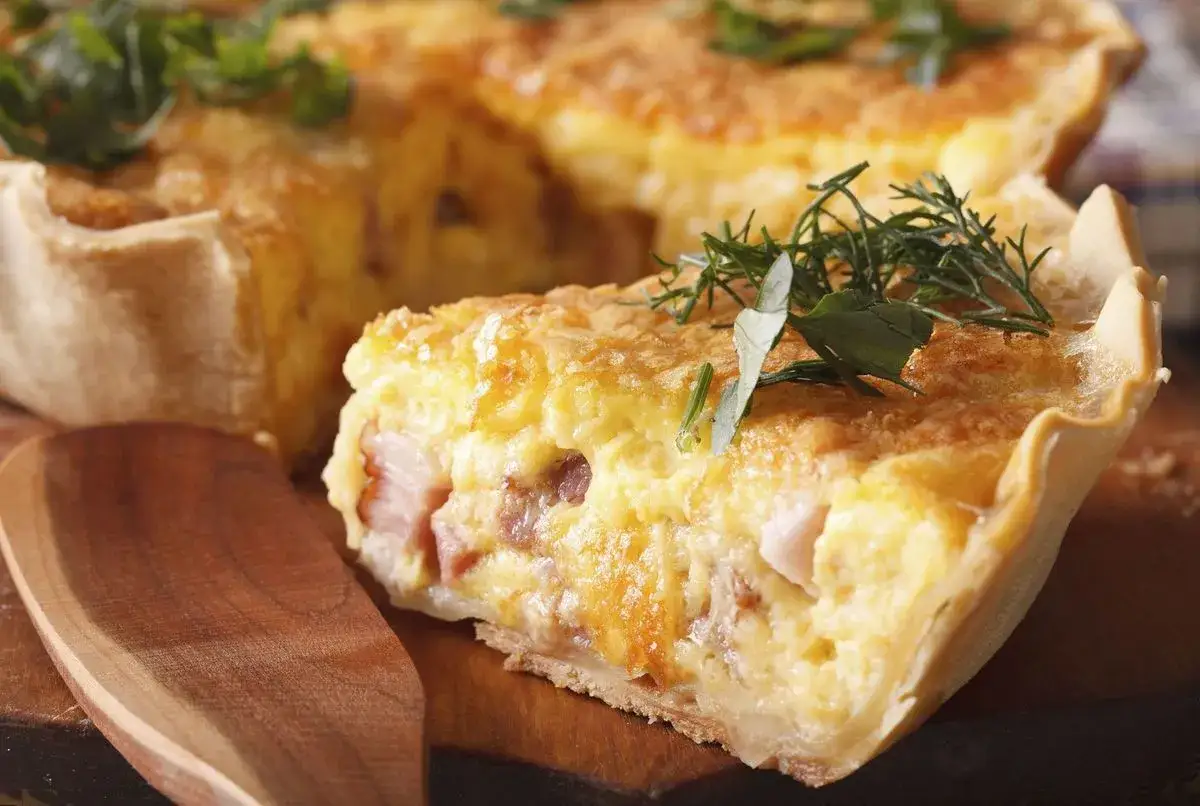 Une part de quiche lorraine, spécialité lorraine, garnie de lardons et d'herbes fraîches, sur une planche en bois avec une cuillère.