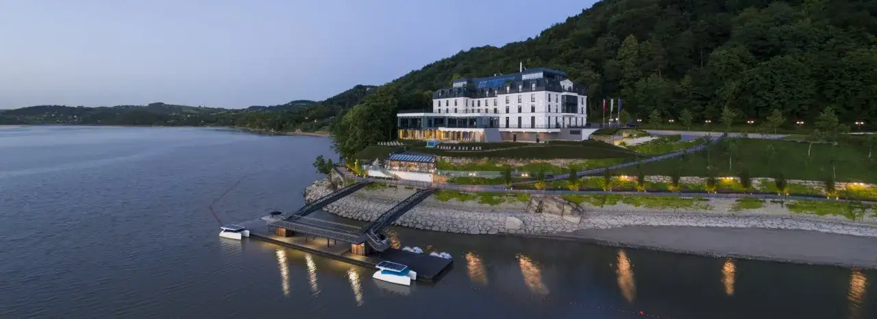 Nowoczesny hotel nad jeziorem Rożnowskim, z pomostem i łódkami, otoczony zielenią.
