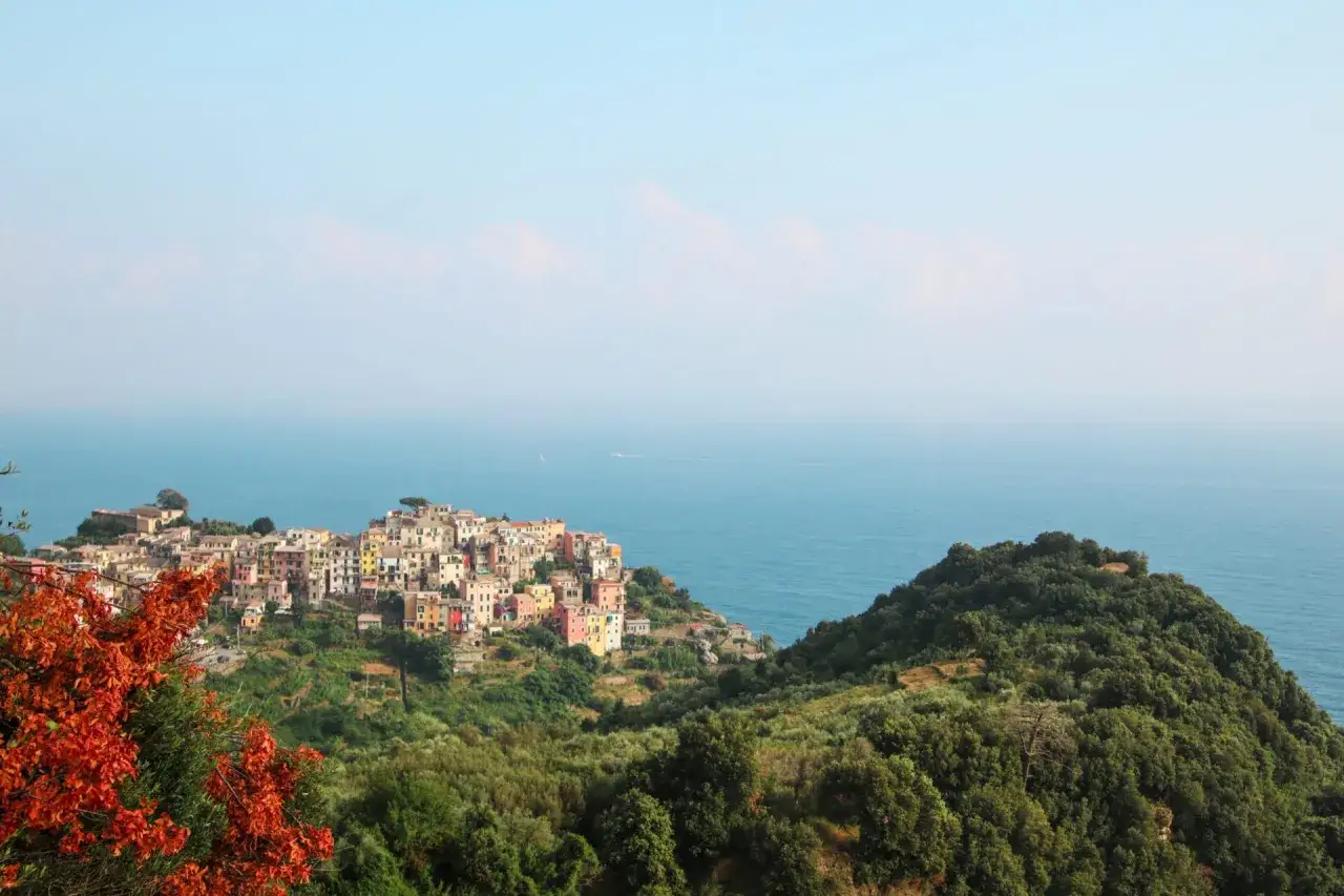 Vue panoramique de **Punta Corvo**, un village coloré accroché à flanc de colline, surplombant la mer bleue. Des arbres verdoyants et des feuilles orangées encadrent la scène.