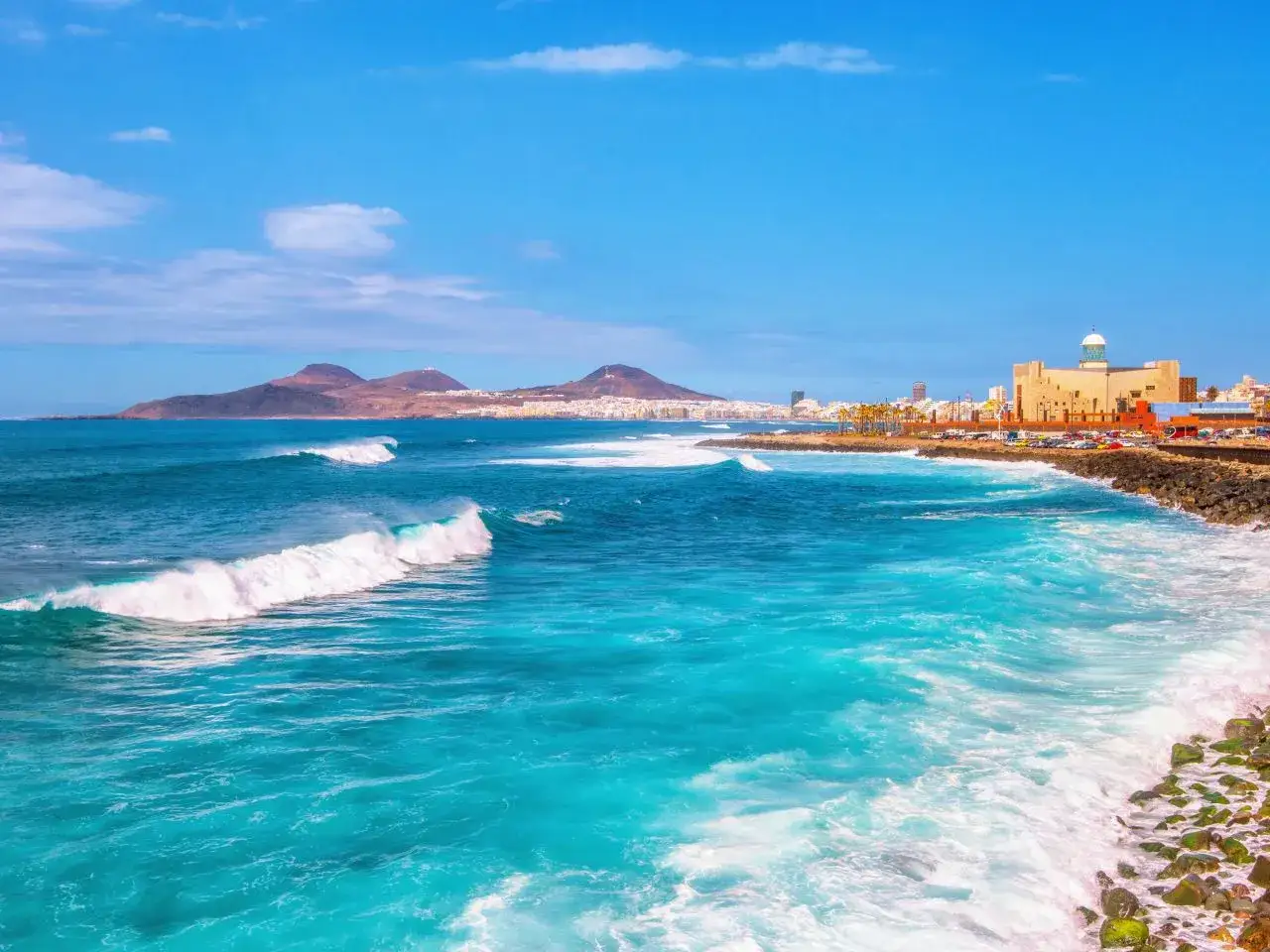 Gran Canaria pogoda czerwiec: turkusowe fale rozbijają się o skalisty brzeg, w tle miasto i góry. Idealny dzień na plażowanie.