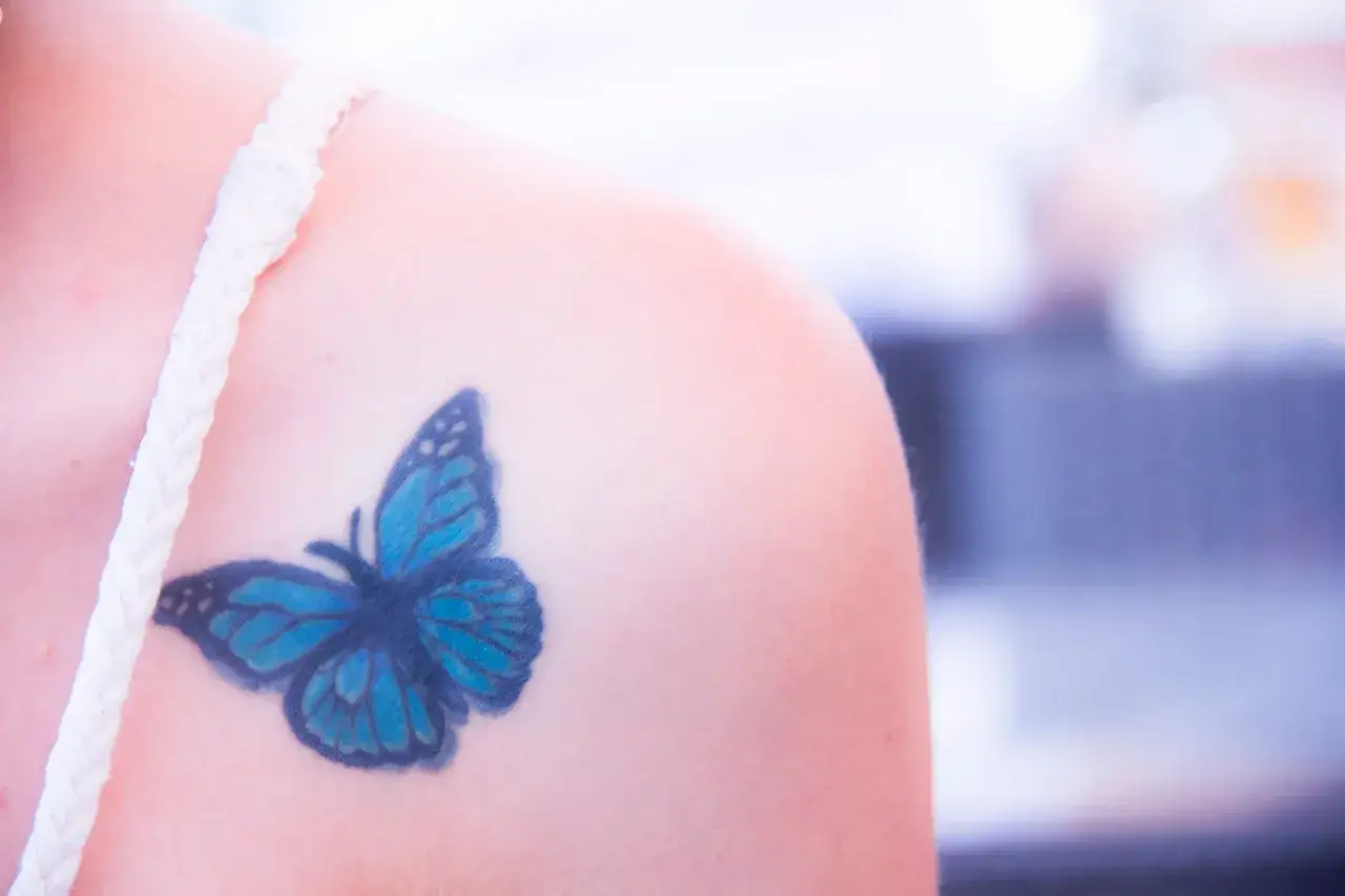 Niebieski tatuaż motyl na ramieniu, ozdobiony białym ramiączkiem.