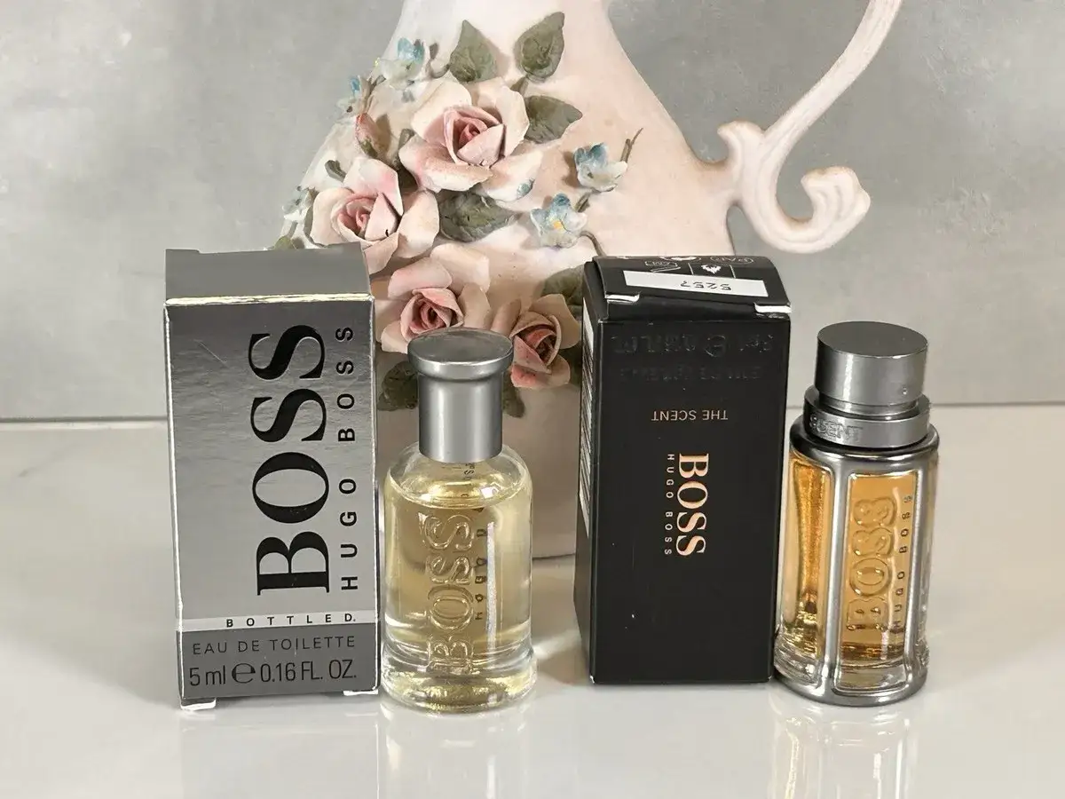 Dwa miniaturowe flakoniki perfum Hugo Boss, jeden z serii Bottled, drugi The Scent, stoją obok swoich pudełek.
